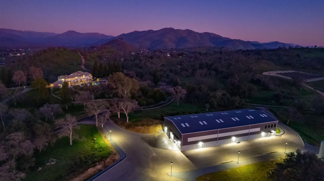 exterior evening shot of custom car warehouse developed by barken near a well lit mansion in the distance near hills