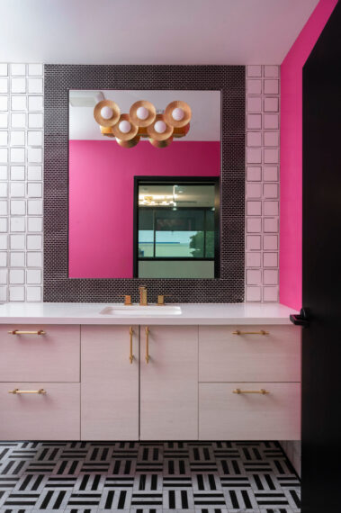Vibrant commercial restroom design with a bright pink accent wall, modern gold vanity lighting, and light wood cabinetry, completed by Barken Construction.