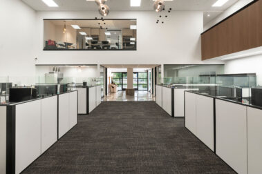 Wide view of a modern open-plan commercial office space with white cubicles and a glass-enclosed upper mezzanine, built by Barken for DV Kap Headquarters.