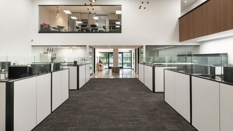 Wide view of a modern open-plan commercial office space with white cubicles and a glass-enclosed upper mezzanine, built by Barken for DV Kap Headquarters.